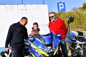 Dziecko siedzi na policyjnym motocyklu. Obok stoi policjant i właściciel Stacji Kontroli Pojazdów.