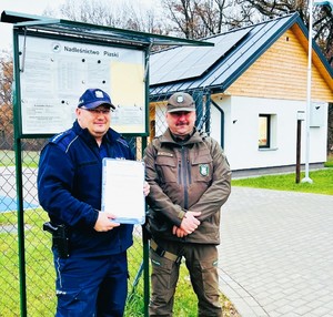 Policjant i strażnik leśny stoją przy tablicy ogłoszeniowej Nadleśnictwa Piaski.