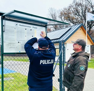 Policjant i strażnik leśny zawieszają ogłoszenie na tablicy ogłoszeń Nadleśnictwa Piaski.