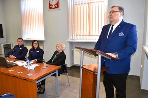 Osoby siedzą za stołem konferencyjnym, jedna osoba stoi przy mównicy.