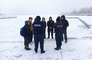 Dwóch policjantów stoi z grupą dzieci na brzegu jeziora.