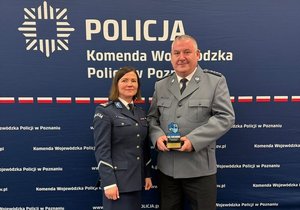 Aspirant sztabowy Krzysztof Tomaszewski z Panią Komendant Komendy Powiatowej Policji w  Śremie - mł. insp. Moniką Szymczak. Oboje w mundurach wyjściowych. Tłem do zdjęcia jest ścianka Komendy Wojewódzkiej Policji w Poznaniu.